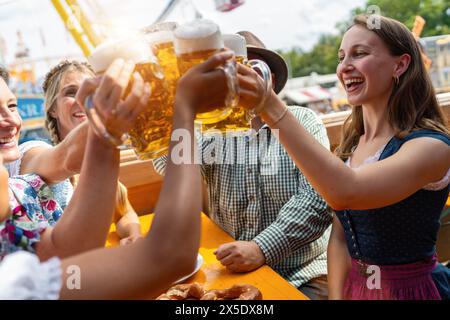Amis joyeux clinking tasses de bière à une table de tente de bière oktoberfest ou dult festival, manèges d'amusement et ciel bleu en arrière-plan Banque D'Images