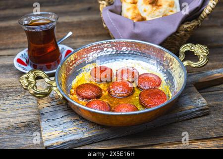 Saucisse turque frite dans une poêle en cuivre, servie avec du thé et du pain, fond en bois, gros plan. Banque D'Images