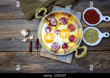 Saucisse turque et œufs frits dans une poêle en cuivre, fond en bois, vue de dessus. Banque D'Images