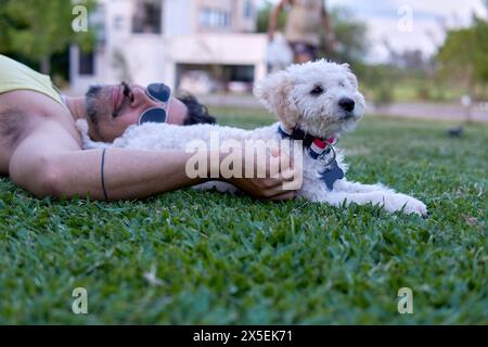 portrait d'homme latin avec des lunettes de soleil couchées sur la pelouse avec son caniche blanc détendu Banque D'Images