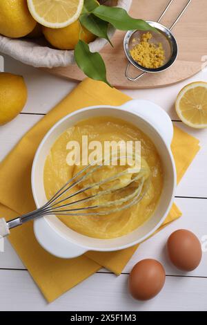 Délicieux caillé de citron dans un bol, ingrédients, fouet et tamis sur une table en bois blanc, plat Banque D'Images