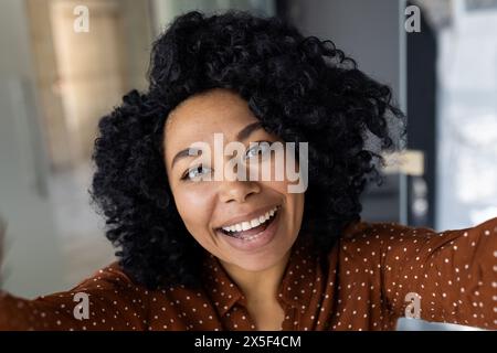 Une jeune femme joyeuse aux cheveux bouclés saisit un moment heureux avec un selfie dans un intérieur élégant et bien éclairé. Son expression est vibrante et engageante. Banque D'Images