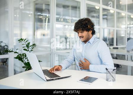 Un jeune homme dans un cadre d'affaires ressent des douleurs abdominales soudaines tout en travaillant sur son ordinateur portable. L'inconfort perturbe sa concentration et son flux de travail. Banque D'Images