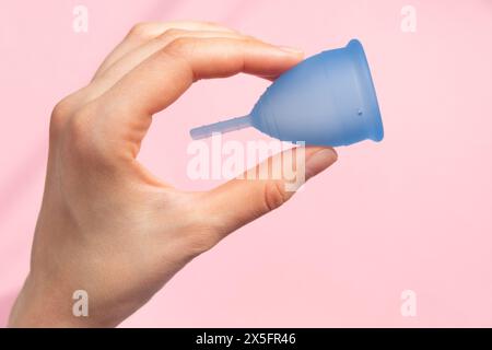 Delicate Woman Hand affiche une coupe menstruelle bleue, placée sur un fond rose doux, symbolisant les solutions d'hygiène menstruelle modernes. Banque D'Images