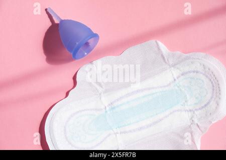 Coupe menstruelle bleue se trouve à côté de la serviette hygiénique sur fond rose vif, illustrant l'approche moderne de l'hygiène féminine. Banque D'Images