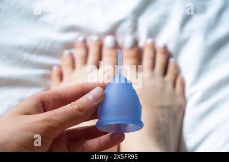 Mains de la femme tenant la coupe menstruelle bleue avec ses pieds en arrière-plan. Le concept de prendre soin de l'environnement, réutilisable. Banque D'Images