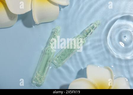 Ampoules de soins de la peau et belles fleurs de plumeria dans l'eau sur fond bleu clair, pose à plat Banque D'Images