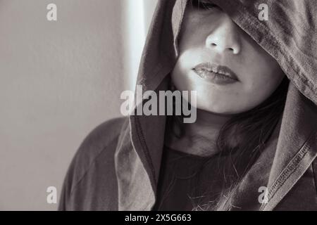Portrait d'une femme mystérieuse dont le visage est partiellement dissimulé par une capuche, capturé en monochrome évocateur, mettant l'accent sur la texture et l'humeur. Banque D'Images
