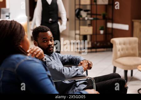 Jeune homme afro-américain en fauteuil roulant parlant avec sa femme en attendant une chambre dans un hôtel adapté aux fauteuils roulants, un homme handicapé arrivant au centre de réadaptation. Hébergement et handicap Banque D'Images