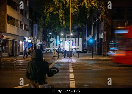 Photo d'un jeune homme à vélo, attendant de traverser une rue où les voitures roulent à Cologne, en Allemagne, la nuit Banque D'Images