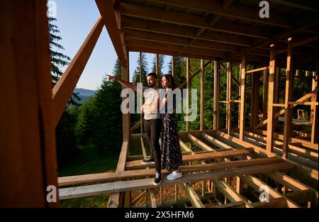 Homme et femme inspectant leur future maison de cadre en bois nichée dans les montagnes près de la forêt. Jeune couple sur le chantier en début de matinée. Concept de construction écologique contemporaine. Banque D'Images