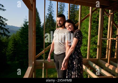 Homme et femme inspectant leur future maison de cadre en bois nichée dans les montagnes près de la forêt. Jeune couple sur le chantier en début de matinée. Concept de construction écologique contemporaine. Banque D'Images