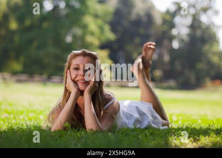 Une femme blonde dans une robe blanche se trouve sur l'herbe verte dans un parc, souriant joyeusement alors qu'elle se détend par une journée ensoleillée. Banque D'Images