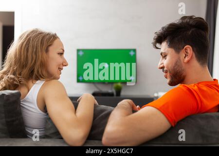 Une jeune femme et un homme sont assis sur un canapé gris, souriant l'un à l'autre, profitant d'un moment détendu et joyeux avec une télévision à écran vert dans le fond Banque D'Images