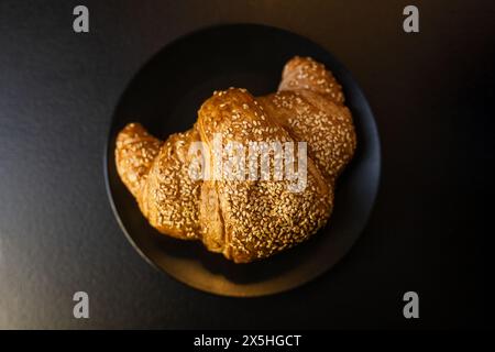 Cette image capture un croissant fraîchement cuit, généreusement recouvert de graines de sésame, présenté sur une assiette sombre et élégante, évoquant un petit déjeuner luxueux Banque D'Images