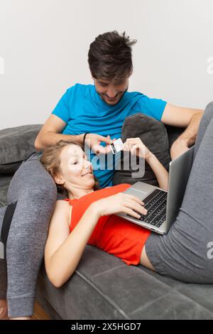 Un jeune couple joyeux se prélasse sur un canapé gris, naviguant sur Internet sur un ordinateur portable tout en discutant d'un achat par carte de crédit. Un scen confortable et quotidien Banque D'Images