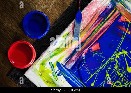 Une vue rapprochée d'une peinture abstraite en cours avec des peintures rouges et bleues vives sur toile, mises en valeur par les pinceaux et les contenants de l'artiste. Banque D'Images