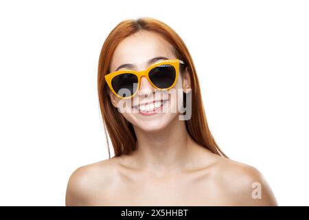 Gros plan d'une jeune femme rousse joyeuse portant des lunettes de soleil jaunes élégantes et montrant un sourire radieux sur fond blanc Banque D'Images