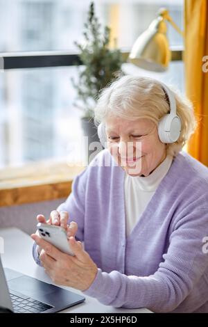 jolie dame âgée assis avec ordinateur portable et smartphone, profitant des technologies modernes. femme âgée souriante, grand-mère utilisant un ordinateur portable seul à la maison, concept de technologie de personnes retraitées. Banque D'Images