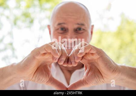 Homme charismatique mature de 65 ans, aîné aimant faisant forme de coeur avec les doigts, la tendresse et les soins, exprimant l'amour et l'affection Banque D'Images