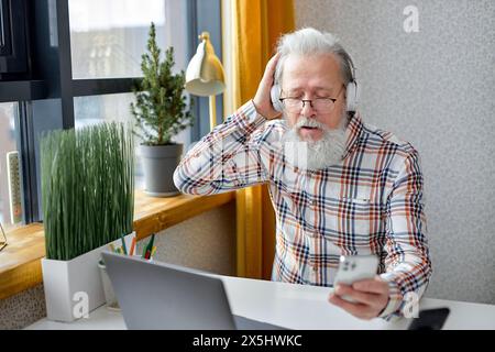 homme âgé assis détendez-vous dans une chambre confortable naviguant sur internet sur ordinateur portable moderne et écouter de la musique à l'aide d'un smartphone, portant un casque. grand-père mûr repos à la maison regarder la vidéo surf sans fil web sur ordinateur portable Banque D'Images