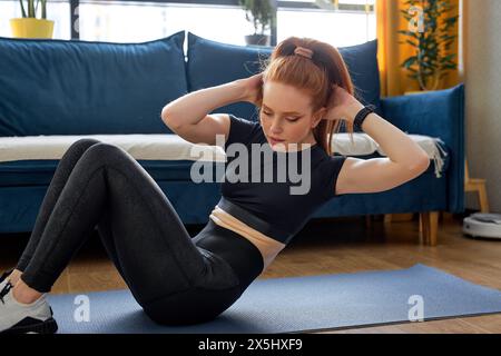 Jeune femme sportive cool attrayante faisant de l'exercice de fitness sur le sol à la maison, portant des vêtements de sport noirs. femme caucasienne rousse engagée dans un mode de vie sain, entraînement seul à l'intérieur Banque D'Images