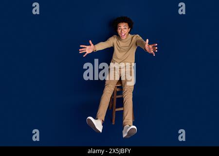 Photo en taille réelle de belle chaise assise de jeune homme tombant porter une tenue beige tendance isolée sur fond de couleur bleu foncé Banque D'Images