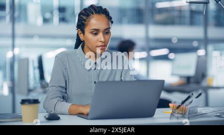 Bureau multi-ethnique : ingénieur INFORMATIQUE professionnel noir travaillant sur le bureau sur ordinateur portable. Femme d'affaires réussie et souriante avec des cheveux tressés fait un travail créatif. Concepteur de produit confiant Banque D'Images
