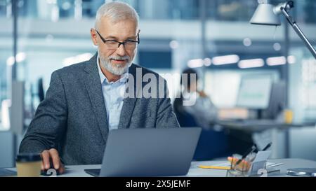 Bureau moderne : Portrait d'un homme d'affaires d'entreprise à barbe d'âge moyen prospère travaillant sur un ordinateur portable à son bureau, souriant. Milieu de travail diversifié avec des professionnels. Banque D'Images