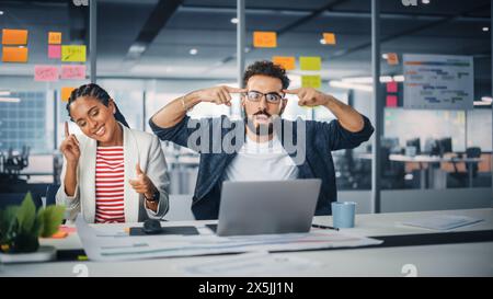 Deux jeunes professionnels heureux au bureau : femme d'affaires noire et homme d'affaires hispanique dansent ensemble, sourire regardant la caméra, assis au bureau. Les spécialistes travaillent dans Creative Office, célèbrent le succès Banque D'Images