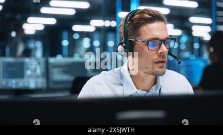 Portrait d'un jeune homme caucasien employé de centre d'appels portant un casque de téléphone parlant à la ligne d'assistance avec un visage sérieux dans un bureau moderne ouvert la nuit. Concept de service d'assistance téléphonique Banque D'Images