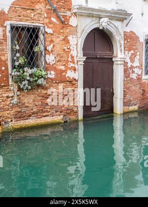 Italie, Venise. Un vieux mur de briques vénitiennes avec du plâtre écaillé le long de l'un des canaux encart avec une fenêtre et une porte tout aussi anciennes. Banque D'Images