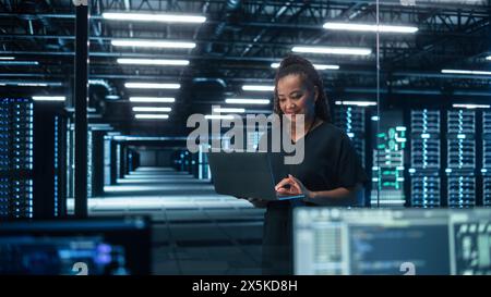 Portrait d'une spécialiste INFORMATIQUE afro-américaine à succès utilisant un ordinateur portable, debout dans un centre de données. L'administrateur système travaille sur les services Web. Cloud Computing, analyse des serveurs, cybersécurité Banque D'Images