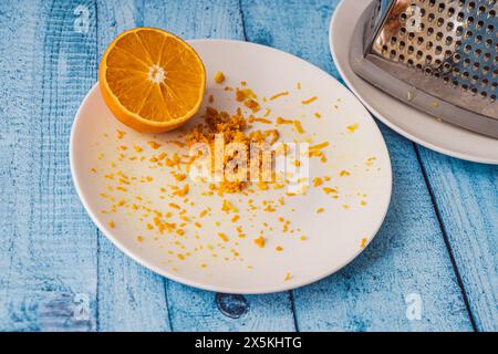 Zeste d'orange râpé sur une plaque blanche sur un plateau en bois avec une orange et une râpe. Recette de crêpe suzette. . Photo de haute qualité Banque D'Images