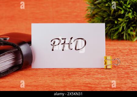 Word PhD. Docteur en philosophie. PHD sur une carte blanche devant un fond orange avec une plante en arrière-plan Banque D'Images