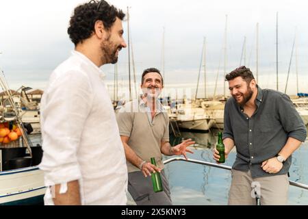 Trois amis riant et socialisant sur un bateau dans une marina, tenant des bouteilles de bière et profitant du moment. La marina et les bateaux créent un dos vibrant Banque D'Images