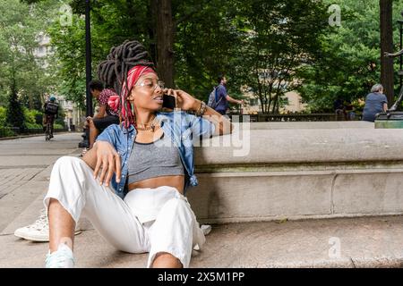 USA, jeune femme assise sur le mur dans le parc, parlant sur un téléphone intelligent Banque D'Images