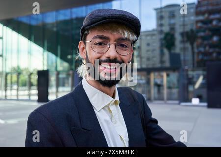 Homme d'affaires hipster regardant la caméra souriant, debout à l'extérieur du lieu de travail. Banque D'Images