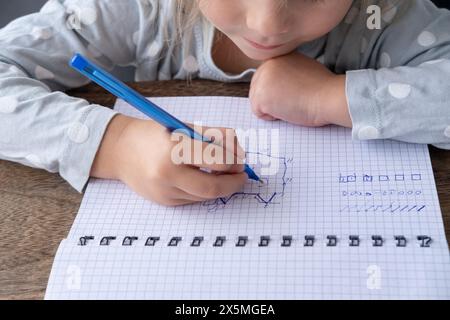 La fille d'âge préscolaire dessine une dictée graphique dans un cahier à carreaux, écrit avant l'école, activités de développement pour les enfants, premiers pas dans l'alphabétisation, C. Banque D'Images