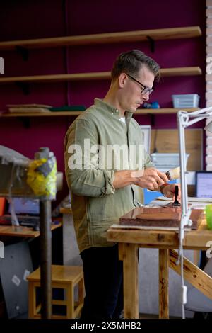 jeune homme beau dans des lunettes perçant des trous dans le cuir avec un poinçon avec un marteau. Outils pour l'artisanat en cuir.gros plan vue latérale Banque D'Images