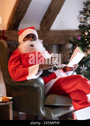 Portrait du Père Noël lisant le livre et buvant du vin dans le fauteuil Banque D'Images