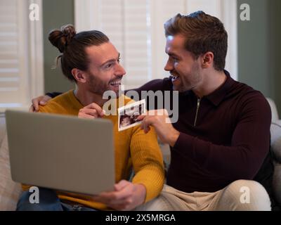 Couple gay montrant l'échographie d'un bébé lors d'un appel vidéo à la maison Banque D'Images