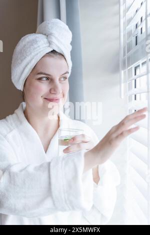 Femme souriante en peignoir et serviette turban regardant par la fenêtre Banque D'Images