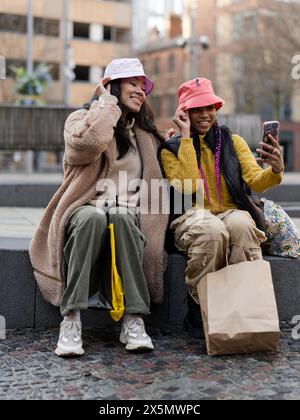 Deux jeunes femmes en milieu urbain, utilisant un smartphone pour prendre des selfies Banque D'Images