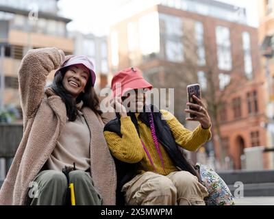 Deux jeunes femmes en milieu urbain, utilisant un smartphone pour prendre des selfies Banque D'Images