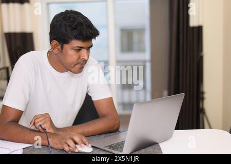 Un Indien se concentre sur son travail tout en utilisant un ordinateur portable dans son salon très éclairé. Banque D'Images