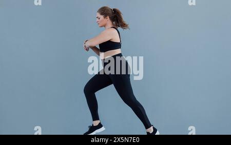 Vue latérale d'une femme grande taille qui court contre un mur gris portant une tenue de fitness noire. Jeune femme avec un jogging de type corps de taille plus et le regardant Banque D'Images
