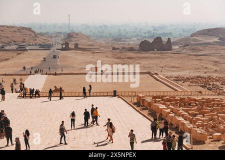 Les touristes traversent la terrasse du temple au temple mortuaire de Hatshepsout à Louxor, avec des ruines désertiques et des terres agricoles verdoyantes faiblement visibles dans le dis brumeux Banque D'Images