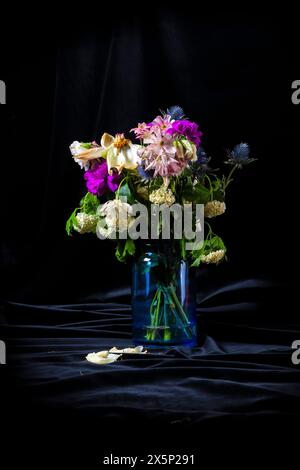 Un vase rempli de fleurs fanées, placé sur un fond noir. Le vase est bleu et contient une variété de fleurs, y compris violettes et blanches. Le Banque D'Images