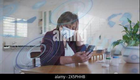 Femme senior biraciale portant un masque, tenant un smartphone à la maison Banque D'Images